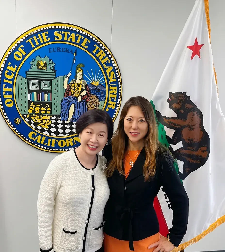 Jackie Wu with California State Treasurer Fiona Ma holding a large check in front of the Office of the State Treasurer seal and the California state flag